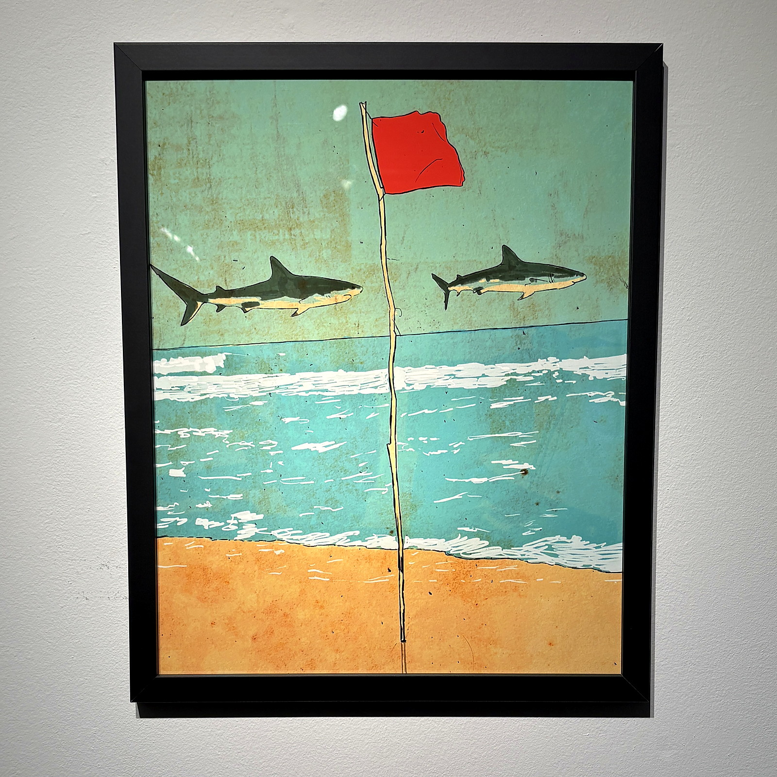 A matte black frame on a white gallery wall: two sharks and a red flag over a beach.